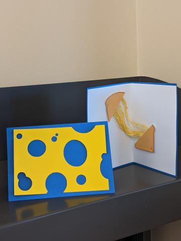 Image of card with a blue background and yellow cheese on the front next to an open card with the image of stretchy grilled cheese.