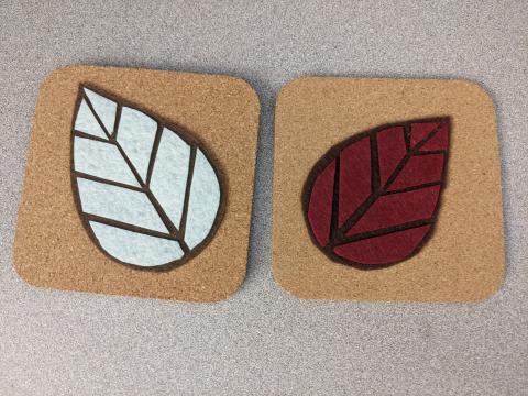 Two cork coaster with felt leaves on them. Left coaster has a leaf with a brown background and white leaf segments, right coaster has a leaf with a brown background and maroon segments.